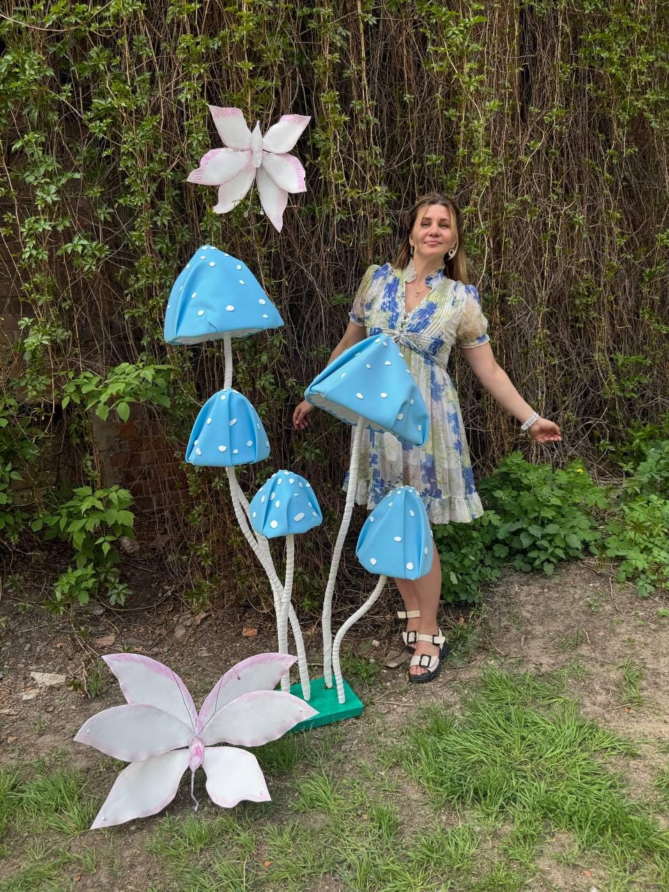 May include: A woman stands in front of a whimsical garden scene with blue and white mushroom sculptures and pink butterfly decorations.