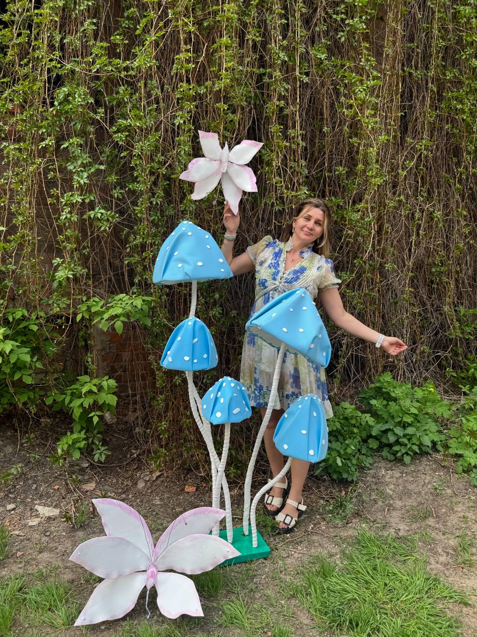May include: A woman stands in front of a backdrop of green vines and holds a large pink paper butterfly.  She is surrounded by four blue paper mushrooms with white stems.  The mushrooms are arranged in a cluster, with the largest mushroom in the center.