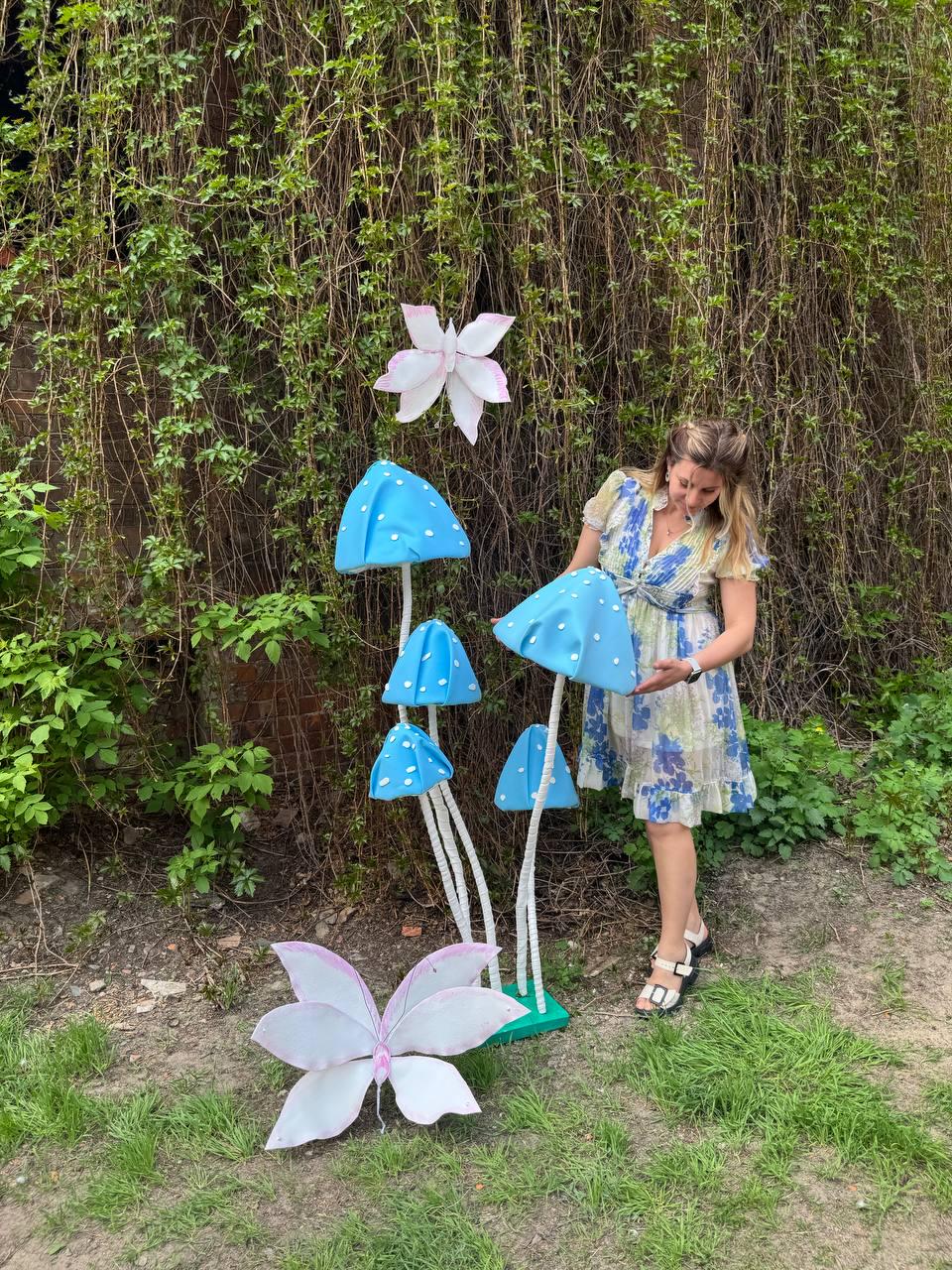 May include: A woman stands in front of a backdrop of green foliage and a cluster of blue and white mushroom-shaped decorations. The mushrooms have white stems and are arranged in a cluster. There are two white butterflies with pink accents on the ground.