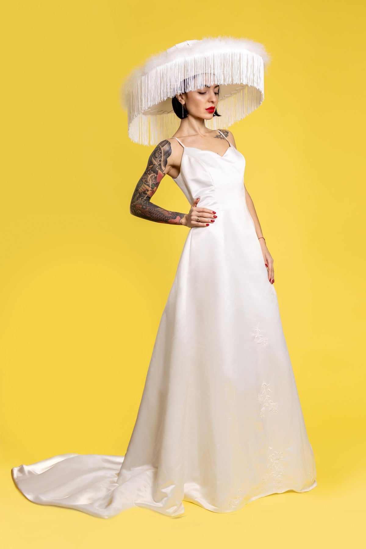 May include: A woman wearing a white wedding dress with a large white feathered hat. The hat has a wide brim and is decorated with fringe. The woman is standing on a yellow background.