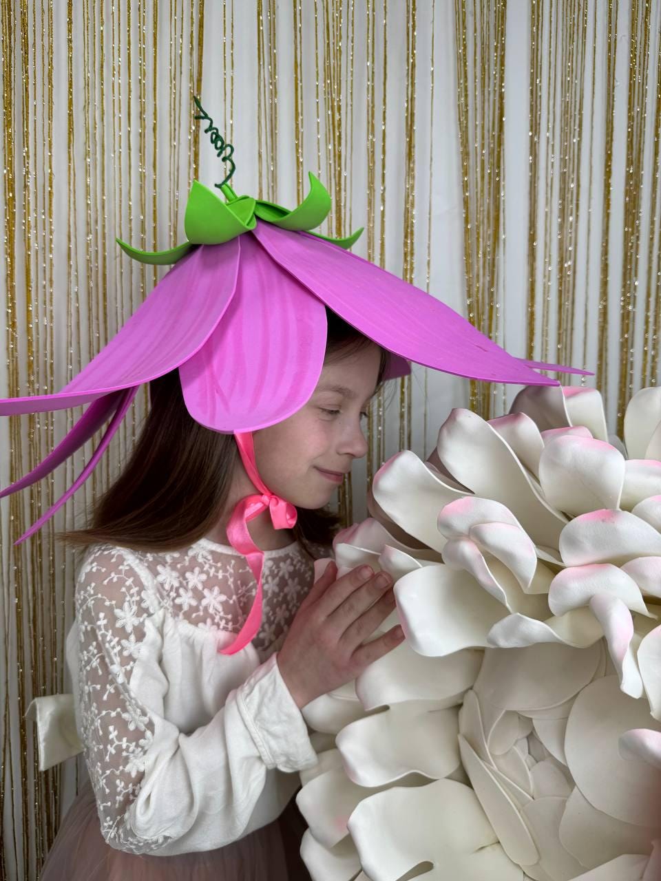May include: A young person wearing a pink flower hat with green leaves and a pink ribbon. They are holding a large white flower.