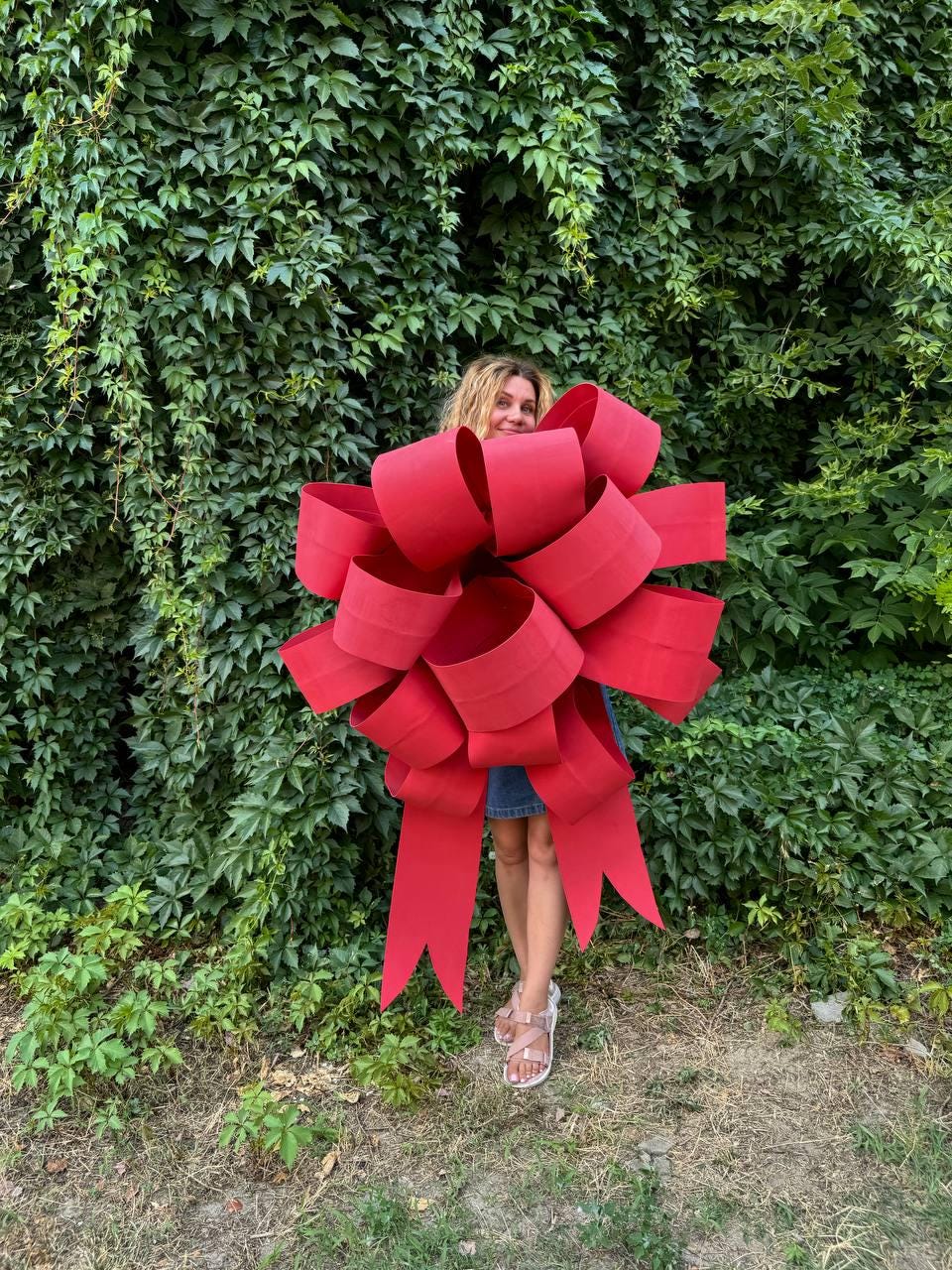 May include: A large, red, oversized bow with long tails. The bow is made of a thick, shiny material and is tied in a classic bow shape. The bow is standing in front of a green leafy wall.