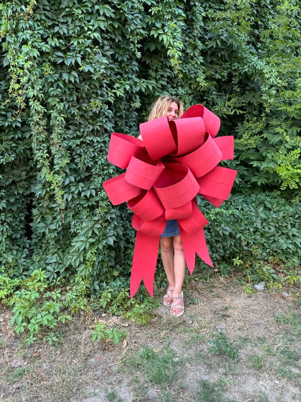 May include: A large, red, oversized bow made of fabric. The bow is being held by a person in front of a green leafy wall.