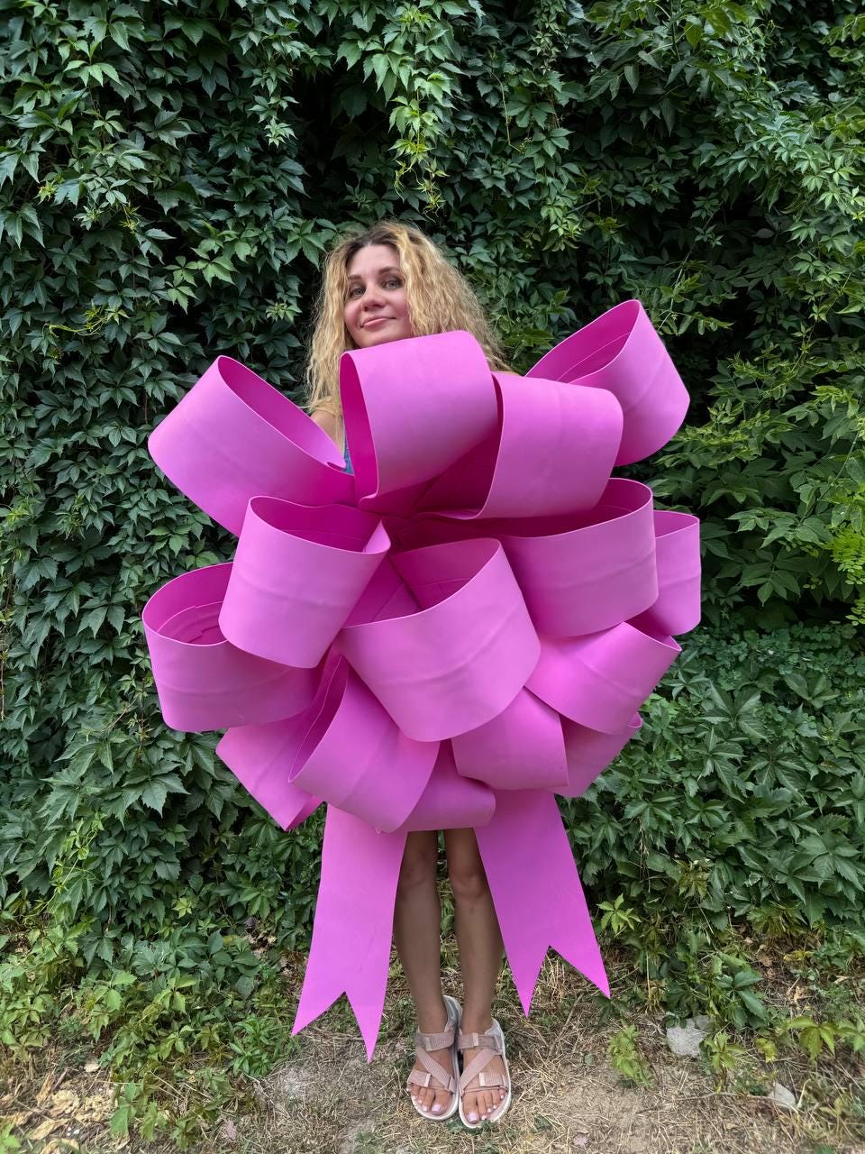 May include: A large pink bow made of fabric, with long tails, is worn as a costume by a person standing in front of a green leafy background.