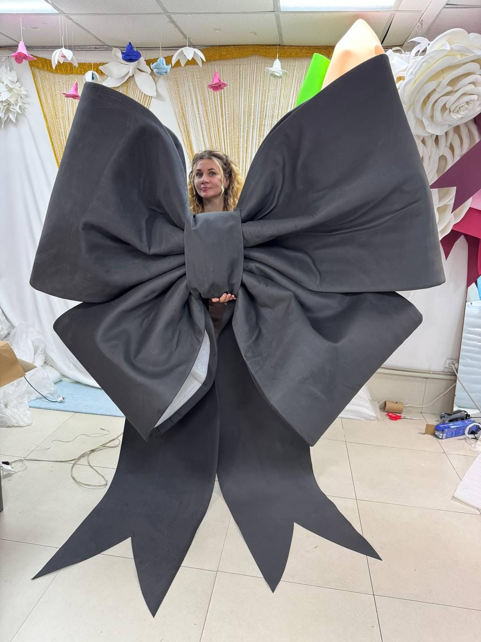 May include: A large black bow made of fabric. The bow is so big that a person is standing inside of it.