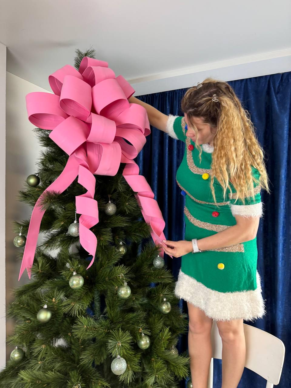 May include: A Christmas tree decorated with silver ornaments and a large pink bow. The tree is being decorated by a person wearing a green dress with white trim and colorful accents. The background includes a blue curtain.