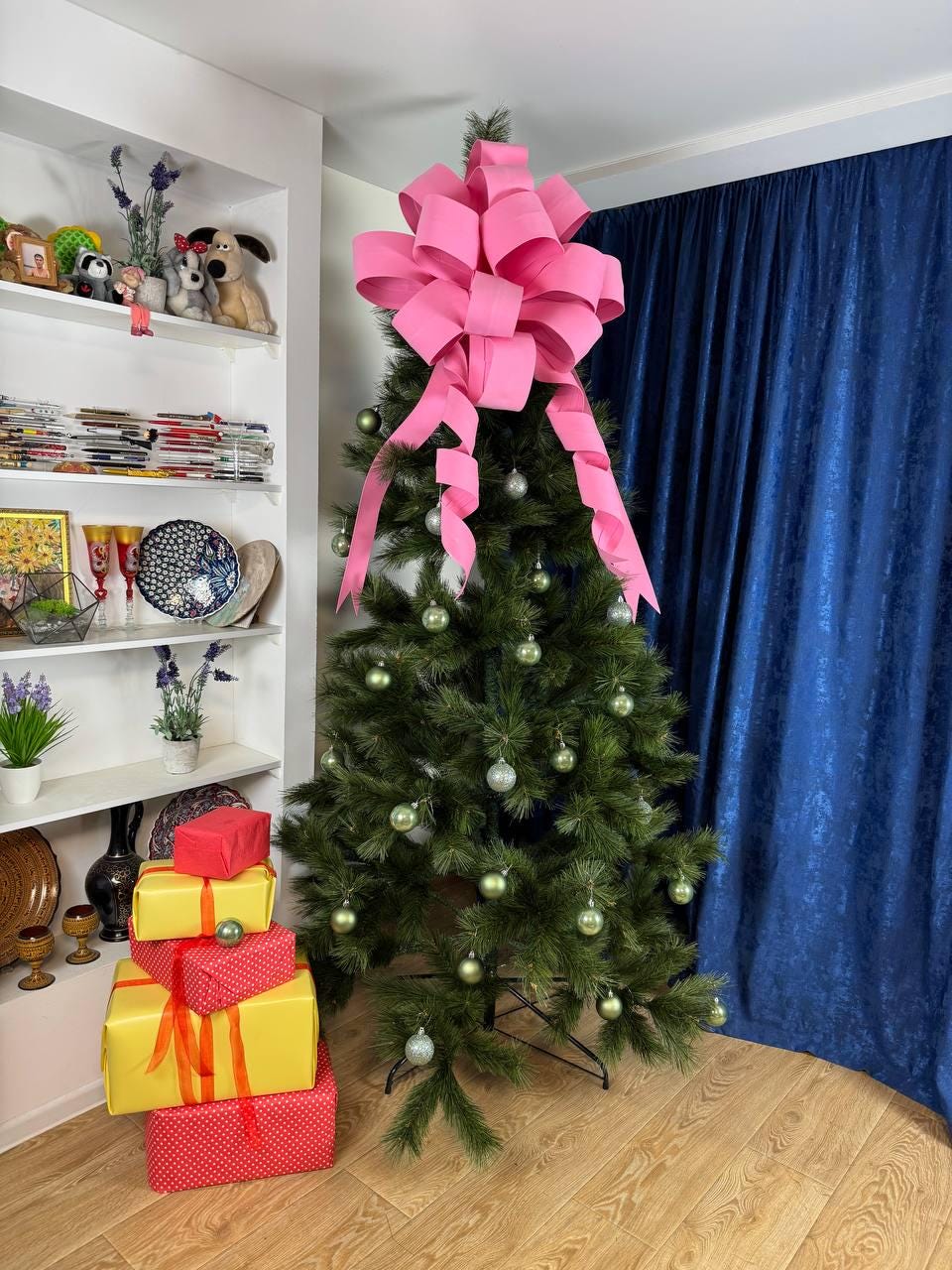 May include: A decorated Christmas tree with a large pink bow on top and green ornaments. Several wrapped gifts in red and yellow are stacked at the base of the tree. The tree is set against a blue curtain.