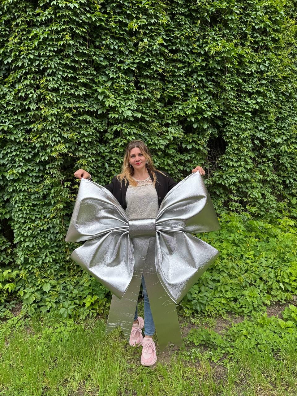 May include: A large, silver metallic bow stands upright in front of a green leafy wall. The bow is oversized, with large loops and a central knot. The bow is made of a shiny, reflective material.