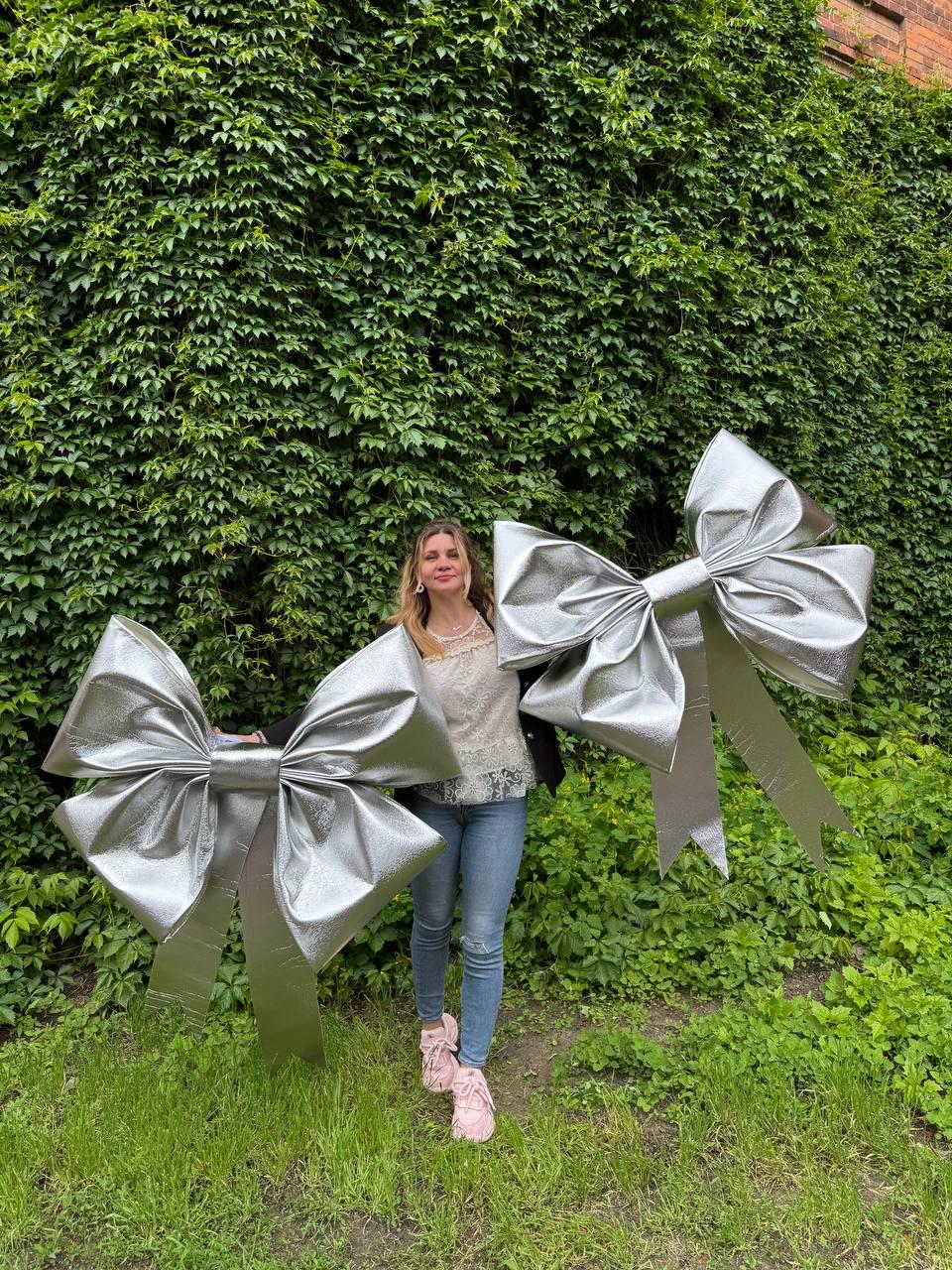 May include: Two large, silver metallic bows with long streamers. The bows are held in front of a person standing in front of a wall of green foliage. The bows have a shiny, reflective surface and are a bright silver color.