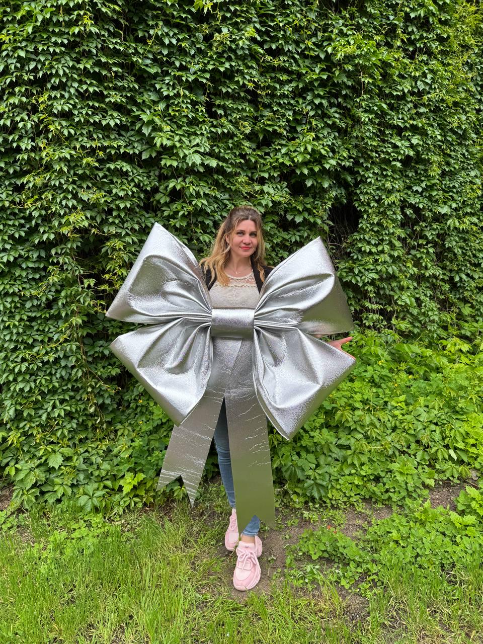 May include: A large, silver bow with long streamers. The bow is made of a shiny, metallic material and is positioned in front of a green leafy background. The bow is large enough to be worn as a costume or decoration.