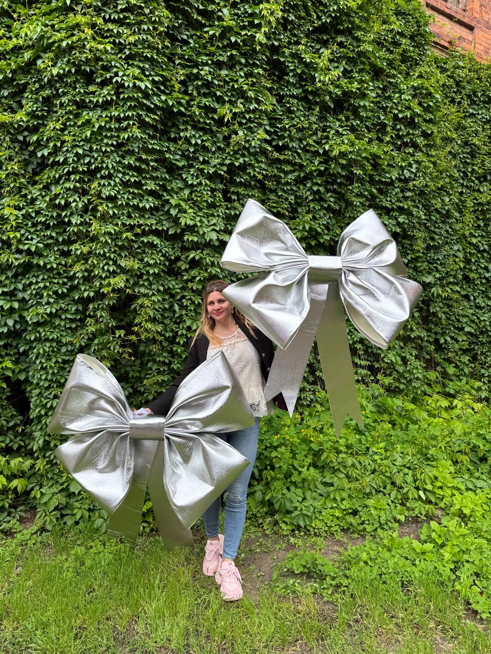 May include: Two large, silver decorative bows. The bows are made of a shiny, metallic material and have a classic bow shape with long streamers. The bows are displayed outdoors against a backdrop of green foliage.