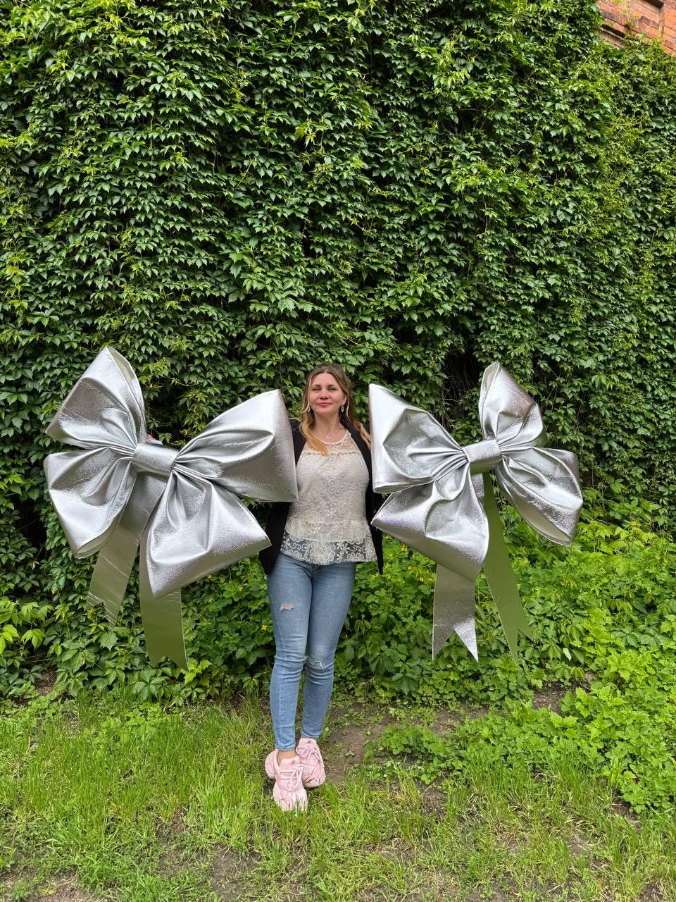 May include: Two large, silver decorative bows with long streamers. The bows are metallic and stand out against a backdrop of green foliage and grass. A person is standing between the bows.