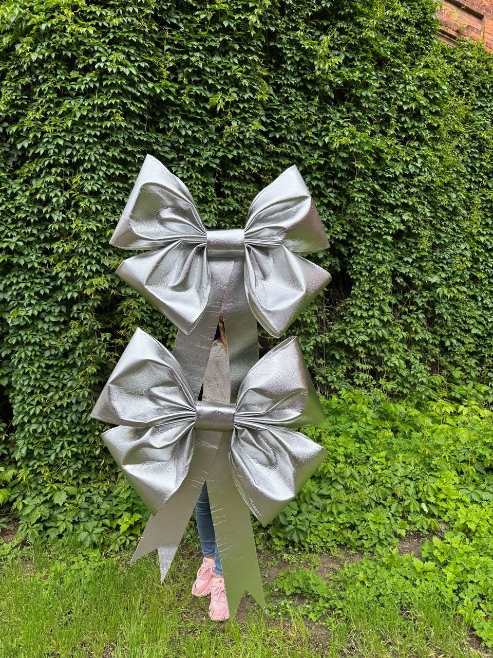 May include: Two large, silver decorative bows with long streamers. The bows are made of a shiny, metallic material and are positioned in front of a lush green wall of foliage. The bows are a bright, eye-catching accent.