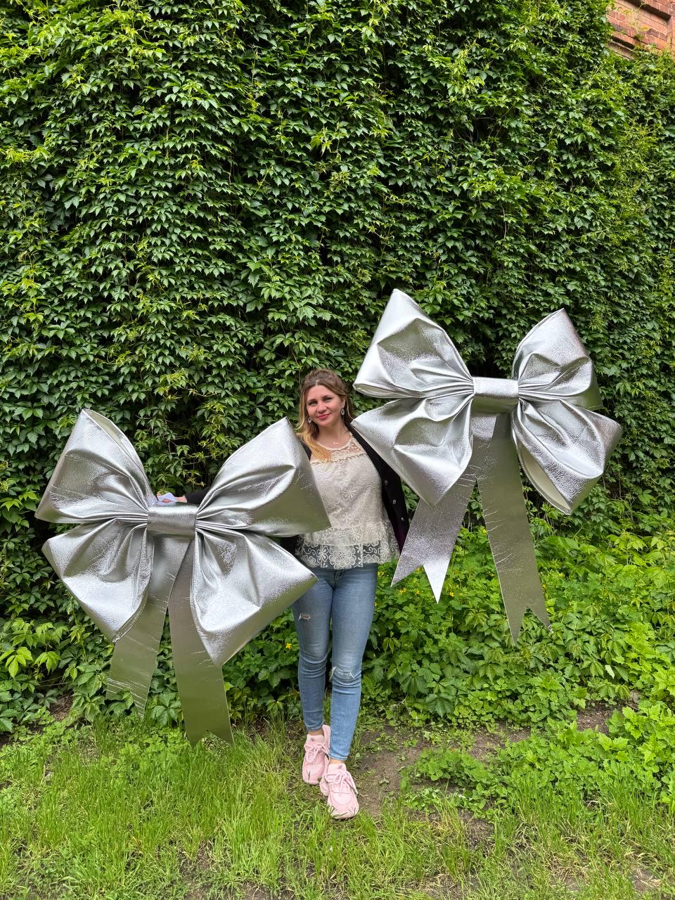 May include: Two large, silver decorative bows with long streamers. The bows are made of a shiny, metallic material and are displayed outdoors against a backdrop of green foliage. The bows are approximately 4 feet wide.