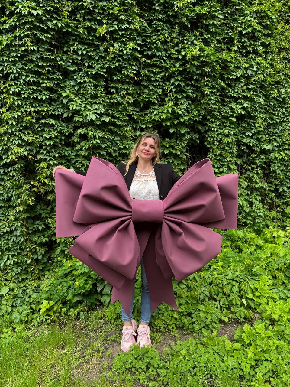 May include: A large, burgundy-colored bow with long streamers. The bow is held in front of a person standing in front of a wall of green foliage. The bow is made of a thick, matte material and has a classic bow shape.