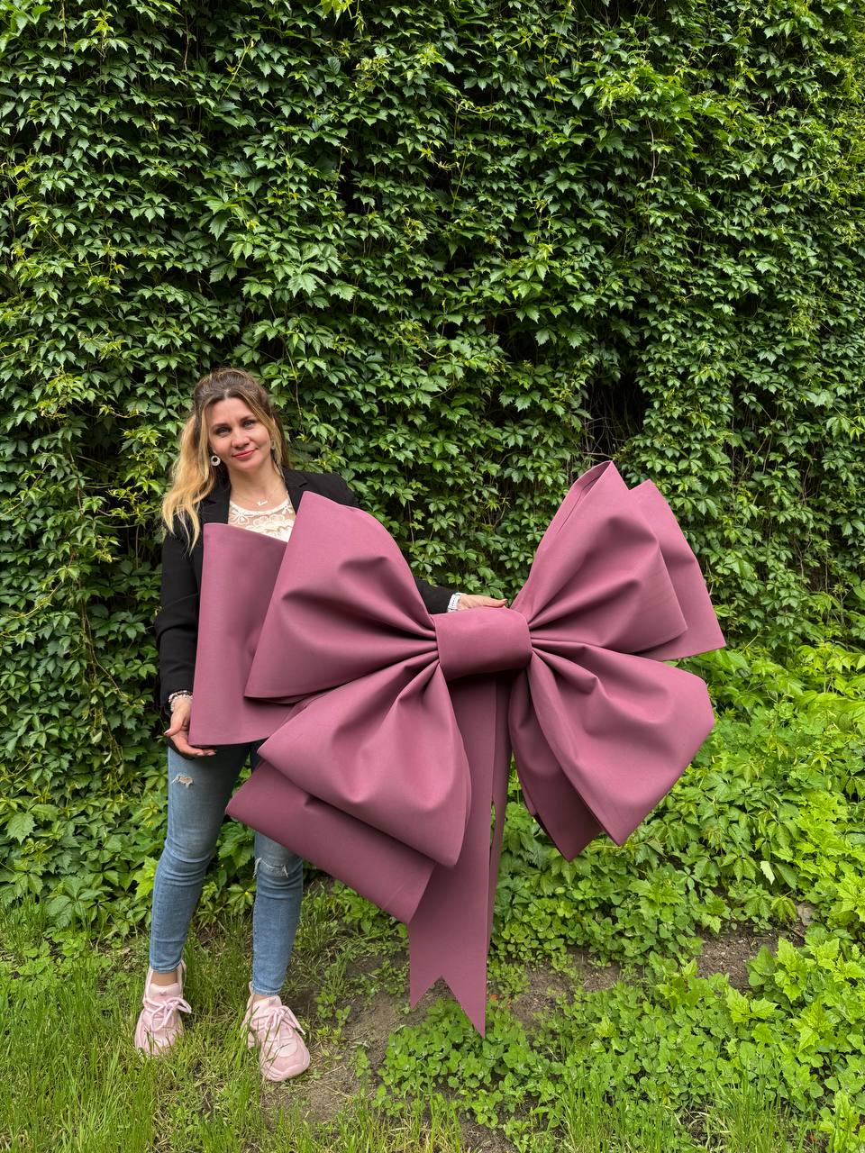 May include: A large, burgundy-colored bow with long streamers. The bow is made of a smooth, matte material and is held in front of a green leafy background. The bow is a decorative item, possibly for a party or event.