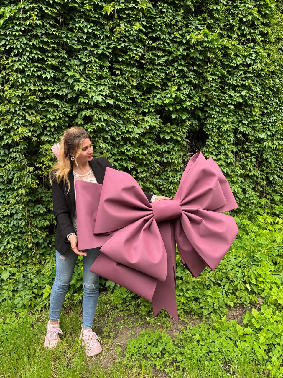 May include: A large, mauve-colored decorative bow. The bow is made of a sturdy material and has multiple loops and tails. The bow is held in front of a green leafy background. The bow is a statement piece.