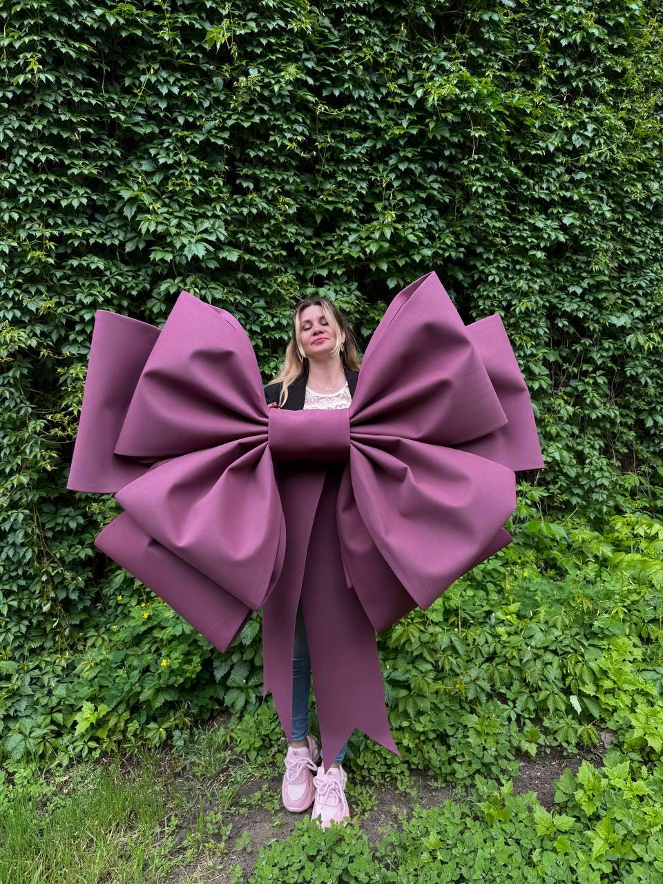 May include: A large, plum-colored bow with oversized loops and long streamers. The bow is the focal point, set against a backdrop of lush green foliage. The bow's color is a deep, rich shade of purple.