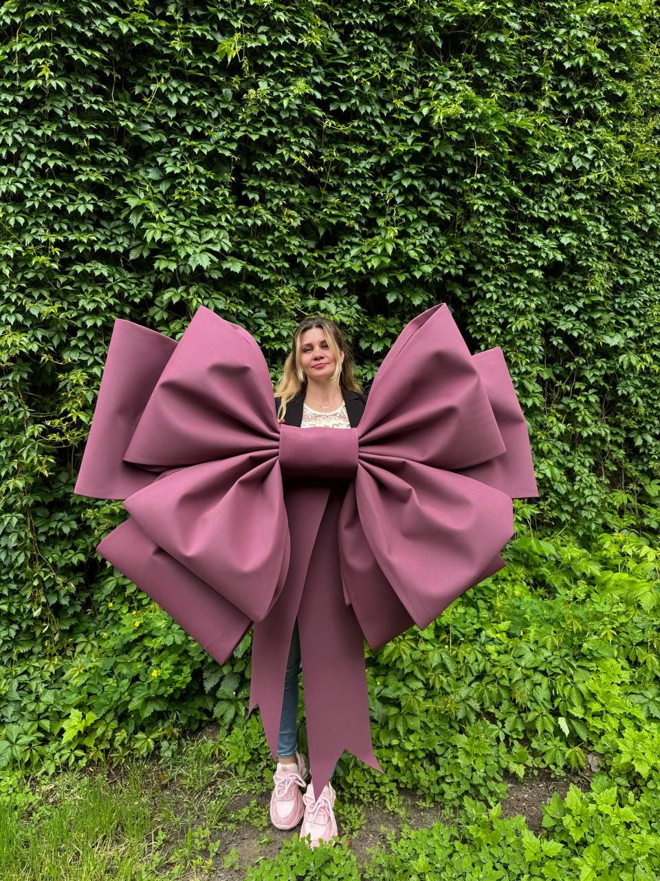 May include: A large, burgundy bow with long streamers. The bow is the focal point, set against a backdrop of lush green foliage. The bow's size and color make it a striking decorative element.