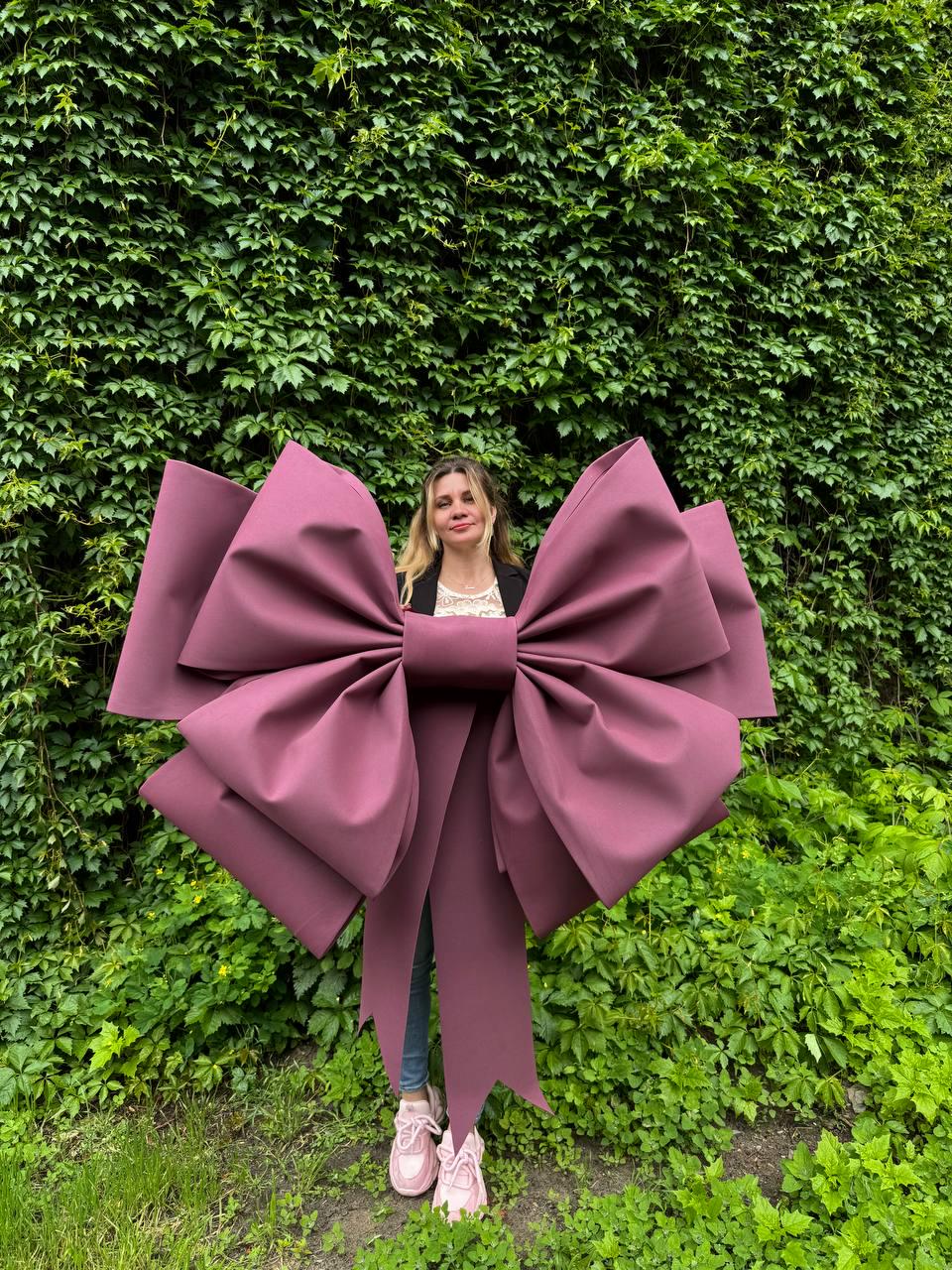 May include: A large, burgundy bow with long streamers. The bow is the focal point, set against a backdrop of lush green foliage. The bow's size and color make it a striking decorative element.