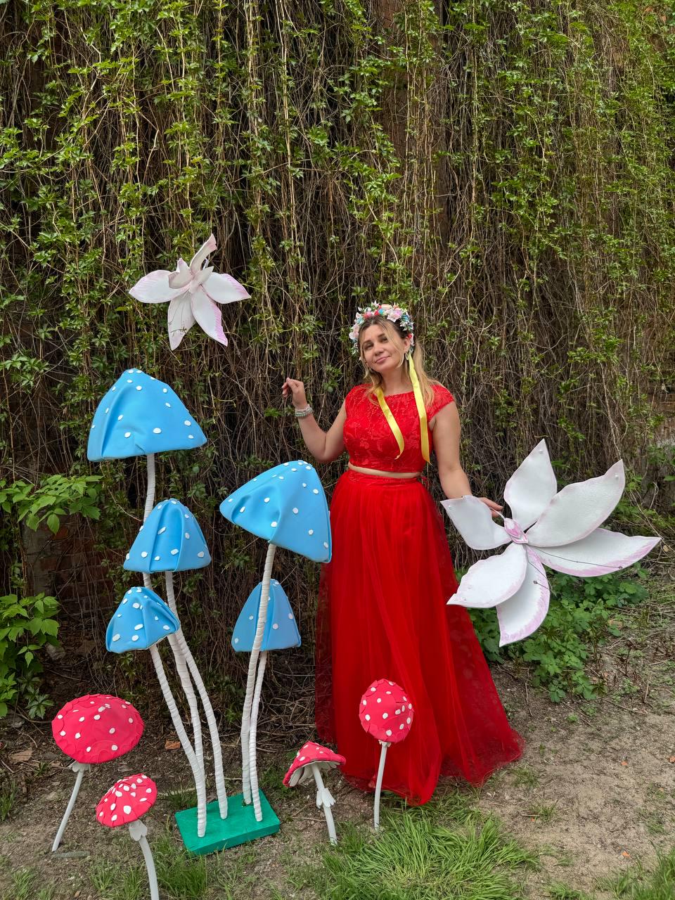 May include: A woman wearing a red dress and a floral crown stands in front of a backdrop of green vines. She is surrounded by large, colorful, paper mache mushrooms. The mushrooms are painted in shades of blue, red, and white.