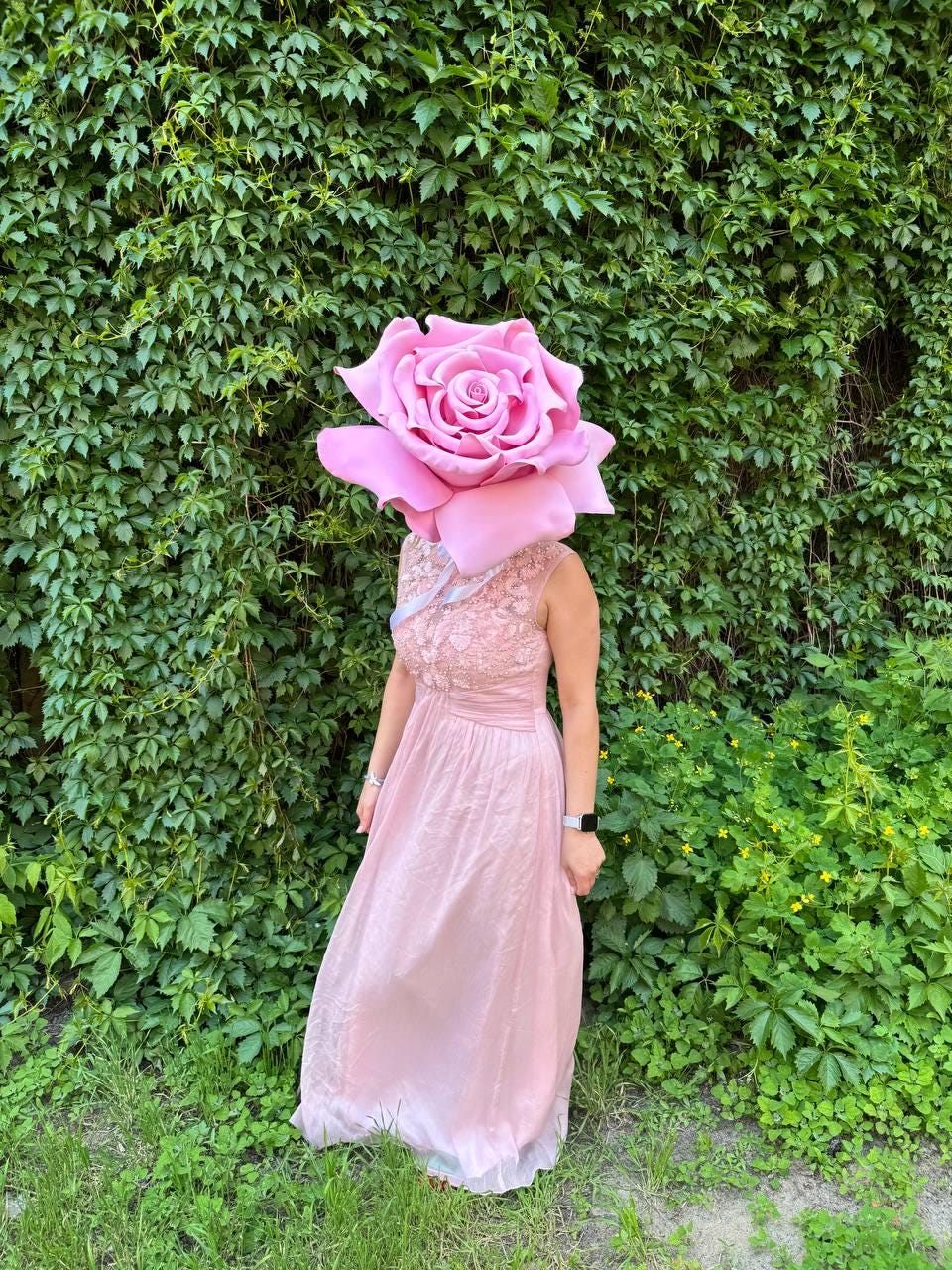 May include: A person wearing a large pink rose headpiece and a pink dress. The dress has a lace bodice and a flowing skirt. The background is a wall of green leaves and grass.
