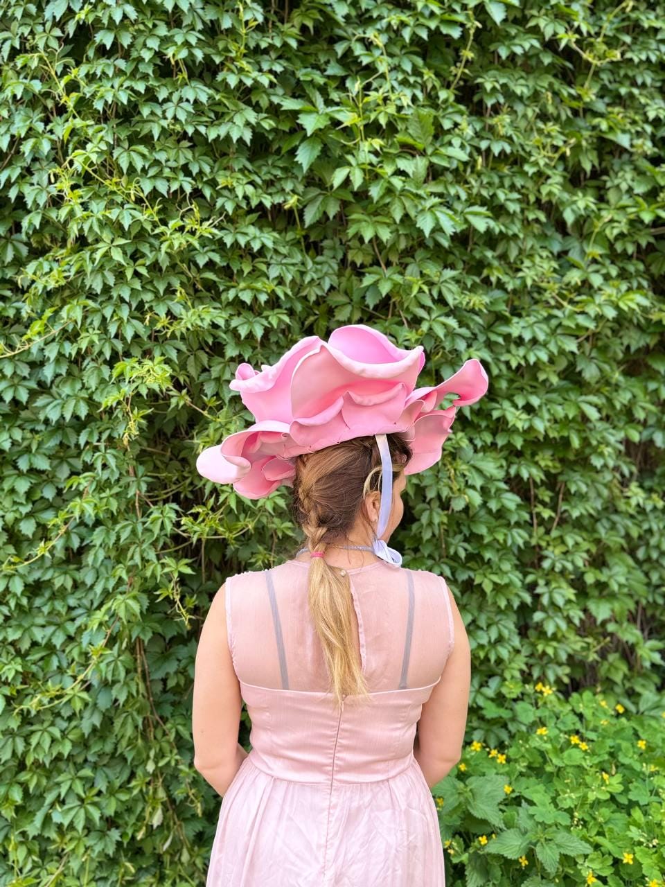 May include: A pink, oversized flower hat with a wide brim. The hat is worn by a person in a pink dress, standing in front of a green ivy wall. The hat's petals are layered and create a dramatic, whimsical effect. The dress has a sheer neckline.