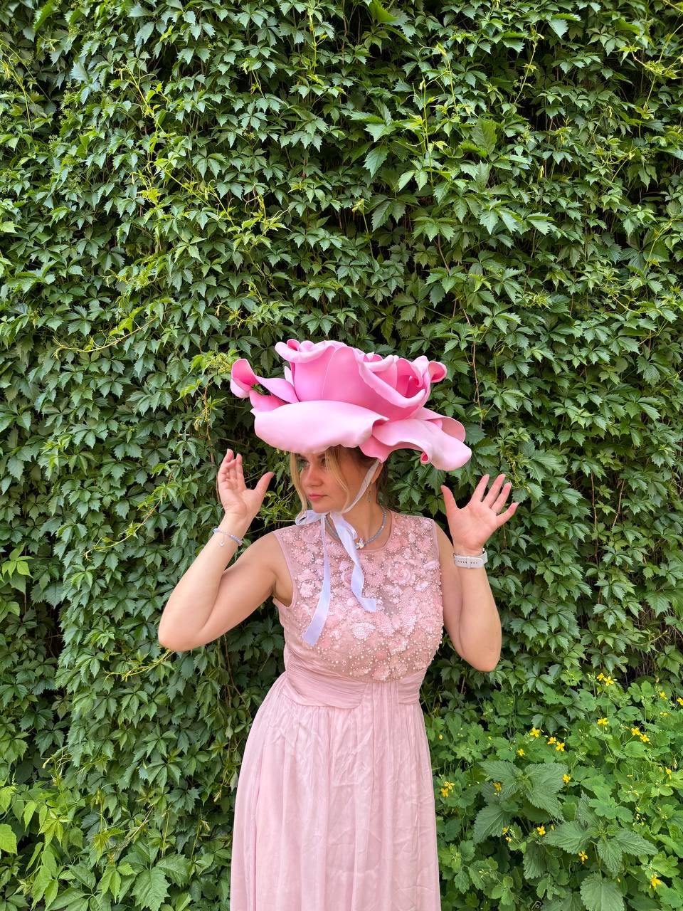 May include: A pink rose-shaped hat with a matching pink dress. The hat is large and detailed, with multiple petals. The dress is sleeveless and has a fitted bodice with floral embellishments. The background is a wall of green leaves.