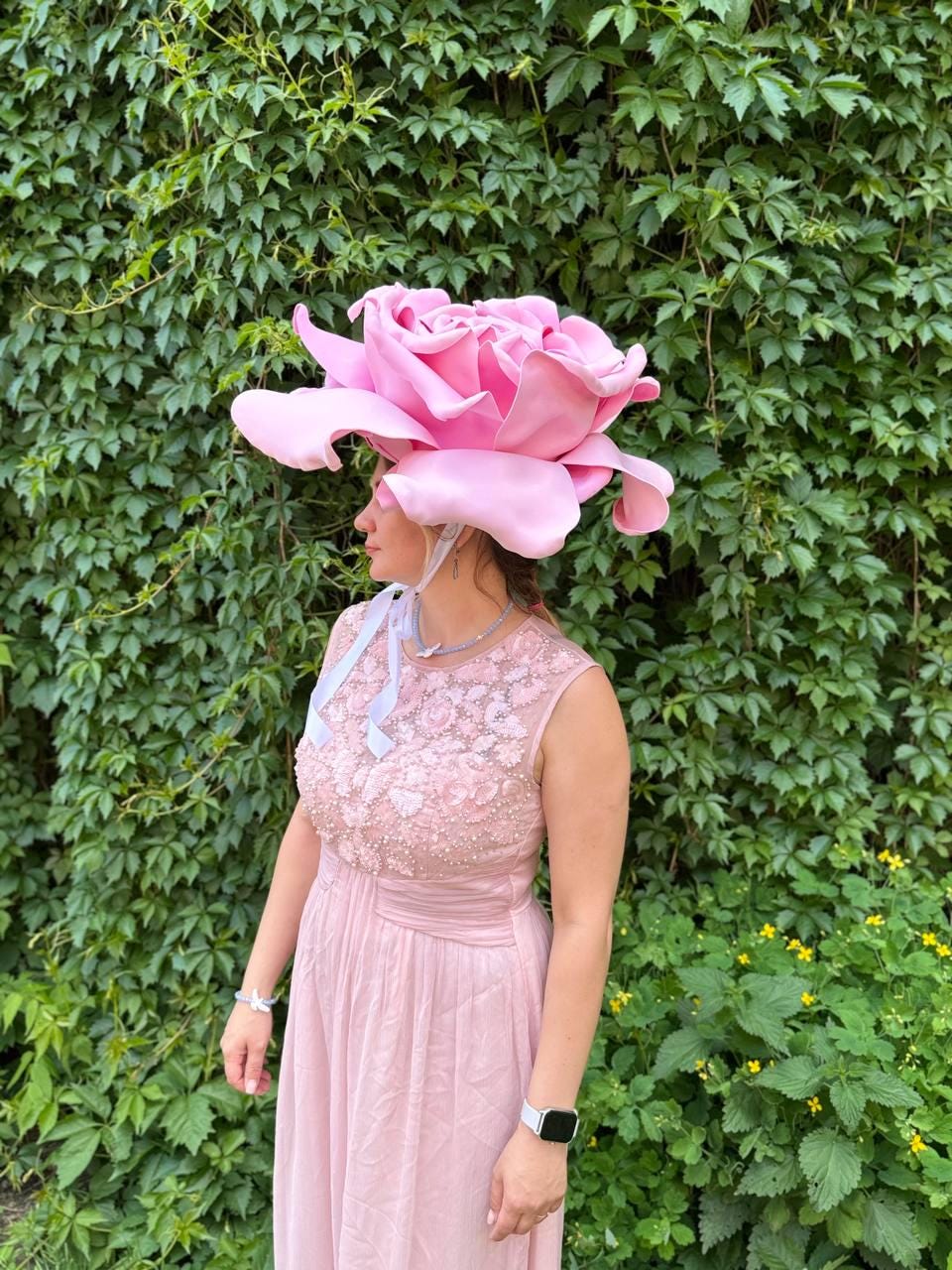 May include: A large, pink rose-shaped hat. The hat is worn with a light pink sleeveless dress. The dress has a beaded bodice and a flowing skirt. A light blue ribbon is tied around the neck. The background is a wall of green leaves.