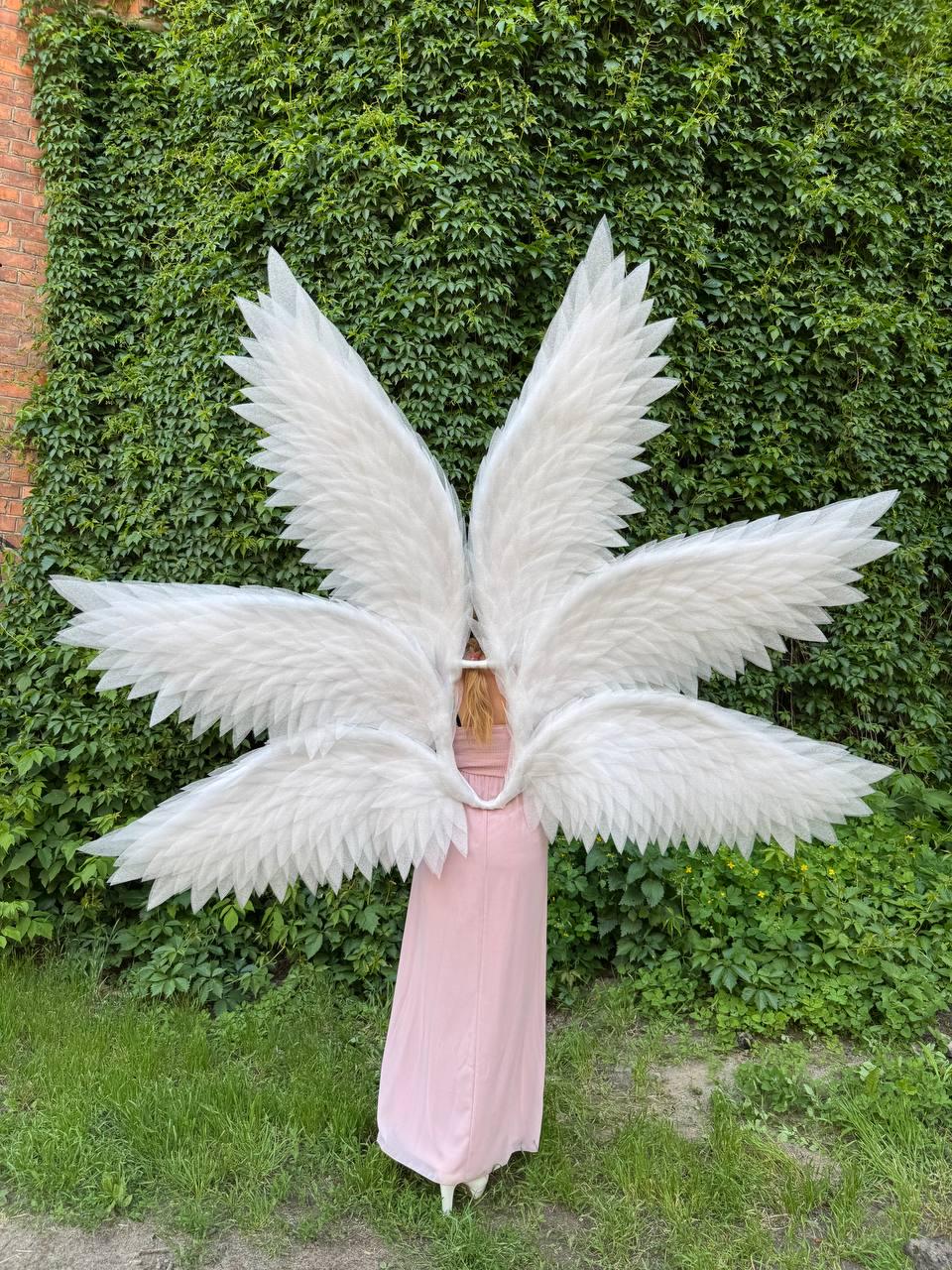 May include: Large, white angel wings with multiple layers of feathers. The wings are worn by a person in a long, light pink dress. The background features a wall covered in green ivy and grass.