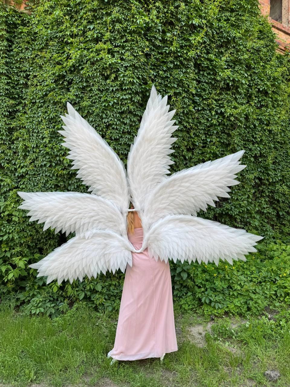 May include: Large, white feathered angel wings with multiple layers of feathers. The wings are worn by a person in a long, light pink dress. The background is a lush green wall of foliage and grass.