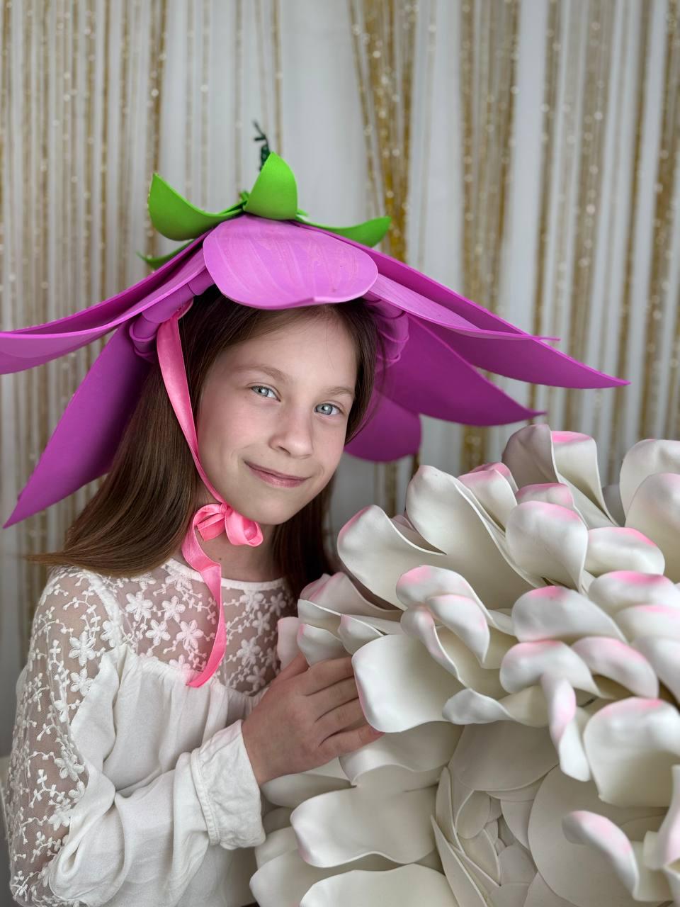 May include: A young girl wearing a pink flower hat with green leaves and a pink ribbon. She is holding a large white flower with pink accents.