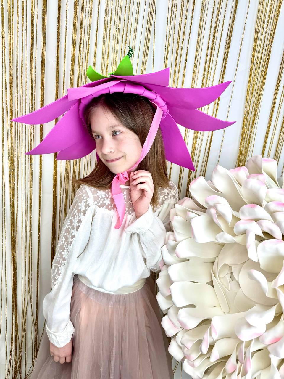 May include: A young girl wearing a pink flower-shaped hat with green leaves. The hat has a pink ribbon tied under her chin. She is wearing a white blouse with lace sleeves and a pink tulle skirt. She is standing in front of a large white flower.