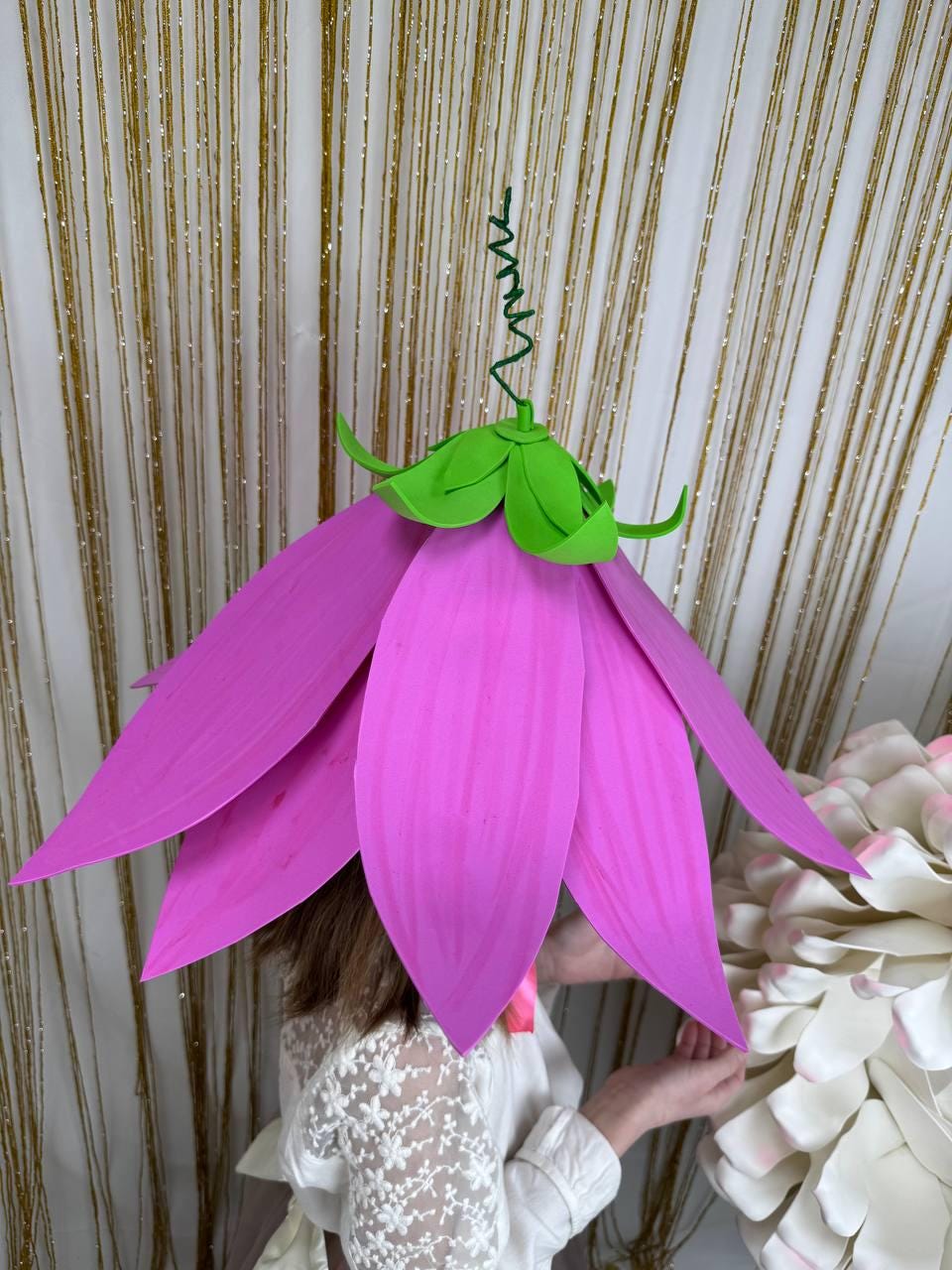 May include: A large pink flower headpiece with green leaves. The flower is made of paper and has a wire stem.