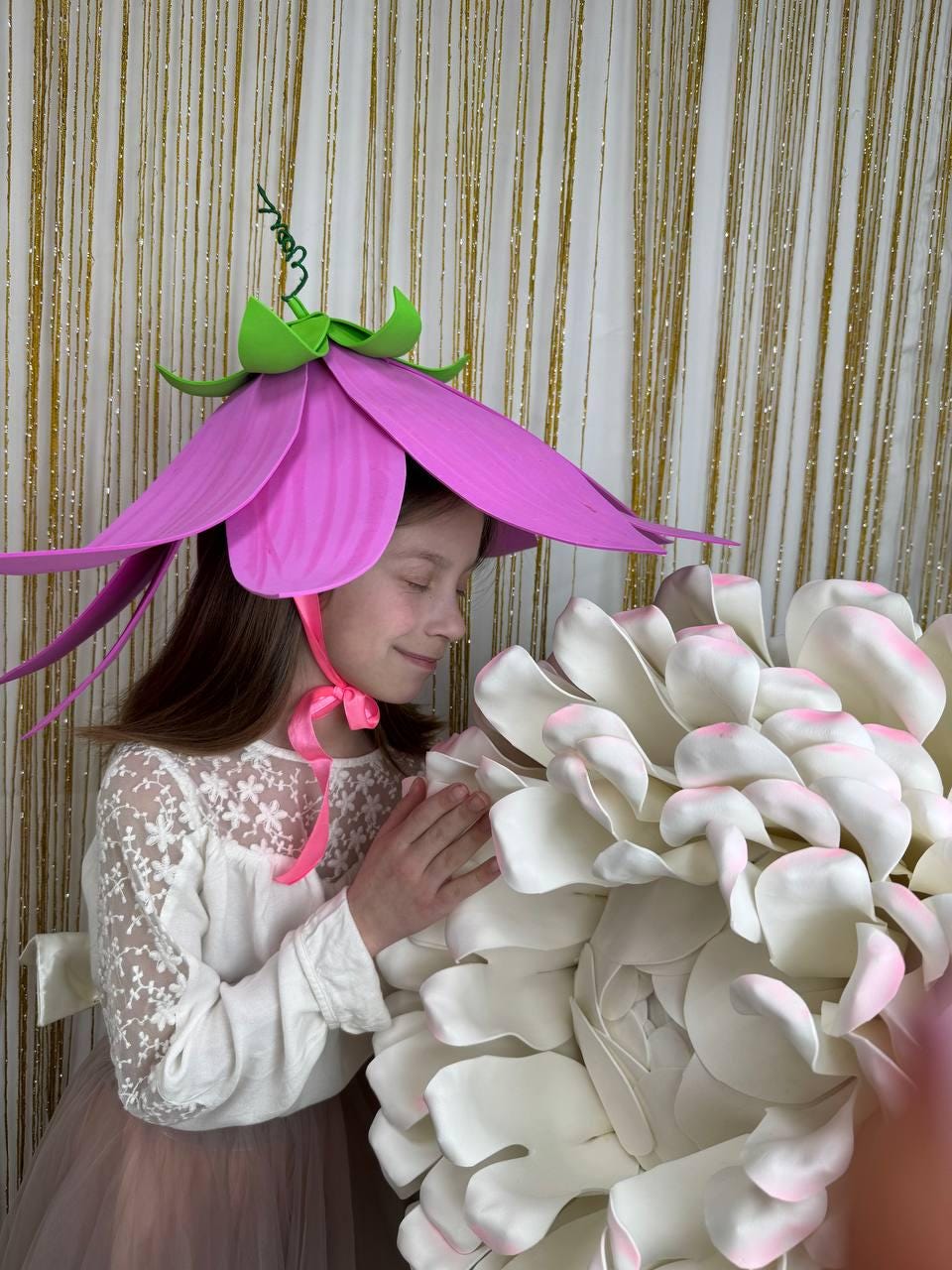 May include: A young girl wearing a pink flower hat with green accents, smiling and holding a large white flower.