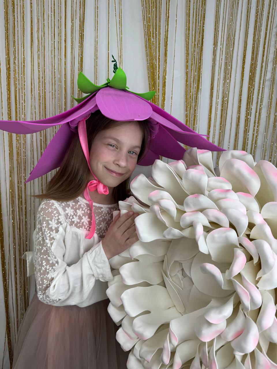 May include: A young girl wearing a purple flower hat with green leaves and a pink ribbon. She is smiling and holding a large white flower with pink accents.