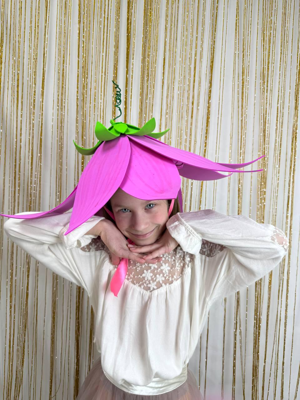 May include: A young person wearing a pink flower hat with green accents. The hat has large petals and a stem. The person is wearing a white blouse with lace trim and a pink ribbon. The background is a gold curtain.
