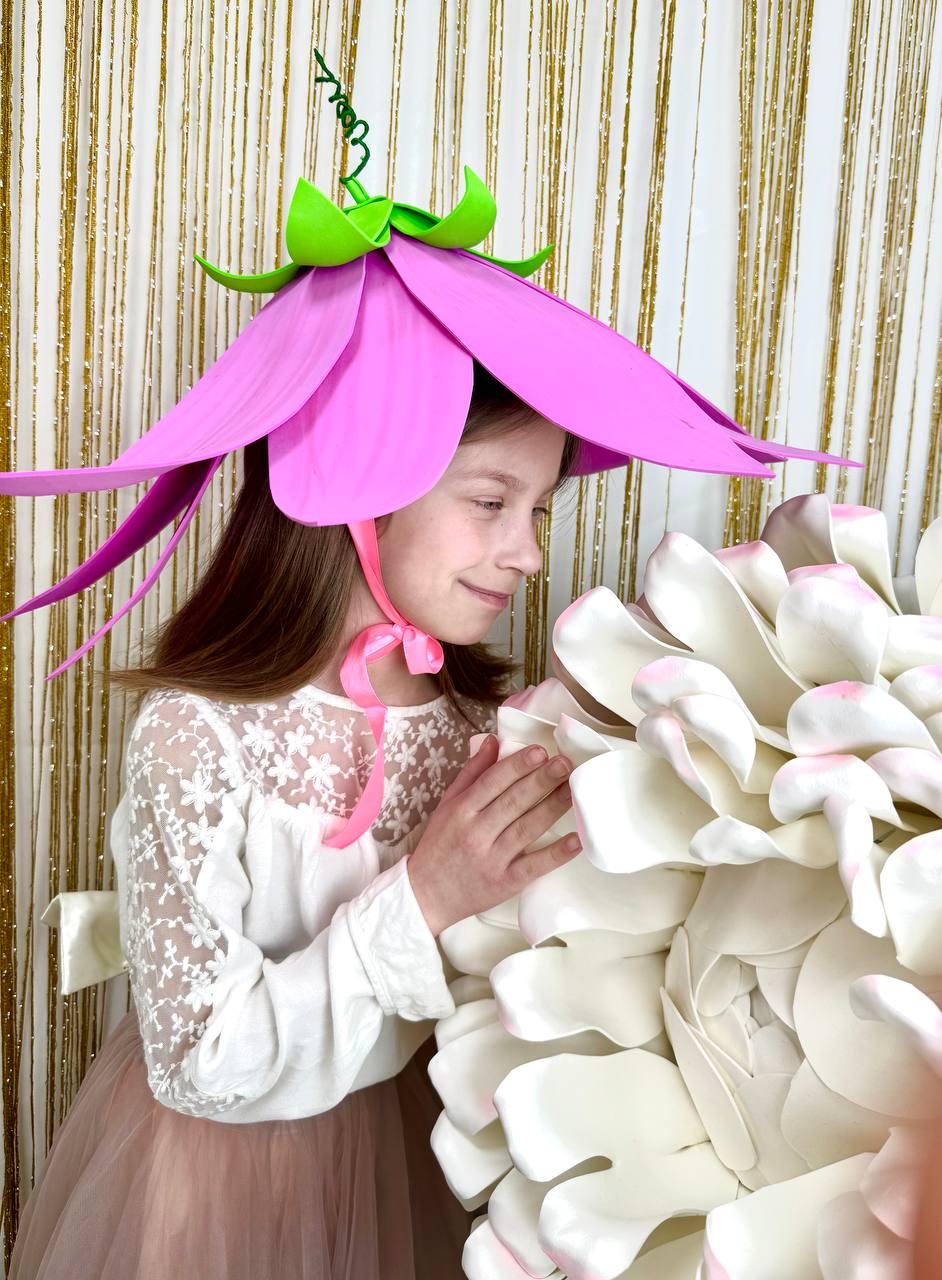 May include: A young girl wearing a pink flower hat with green accents and a pink ribbon. She is holding a large white flower.