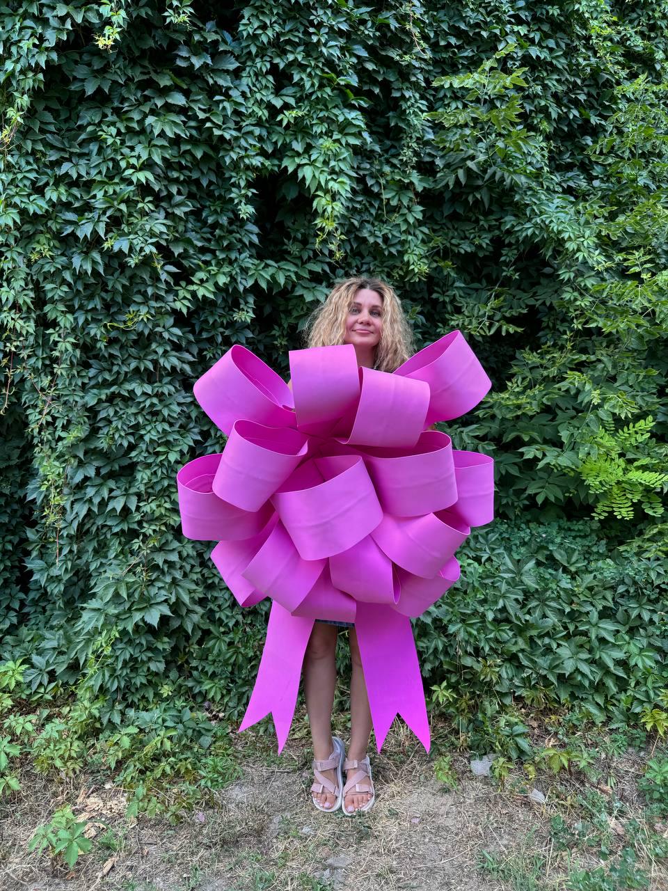May include: A woman stands in front of a green ivy wall wearing a large pink bow. The bow is made of multiple layers of pink ribbon and has long tails.