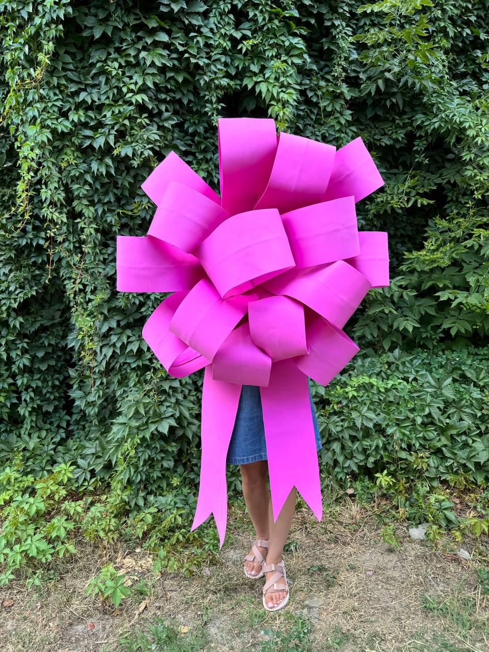 May include: A large, bright pink bow made of paper. The bow is being held in front of a person wearing a denim skirt.