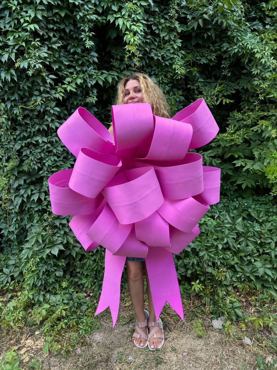 May include: A large pink bow made of multiple layers of ribbon. The bow is large enough to cover a person's torso. The bow is tied with a long ribbon that trails behind the person.