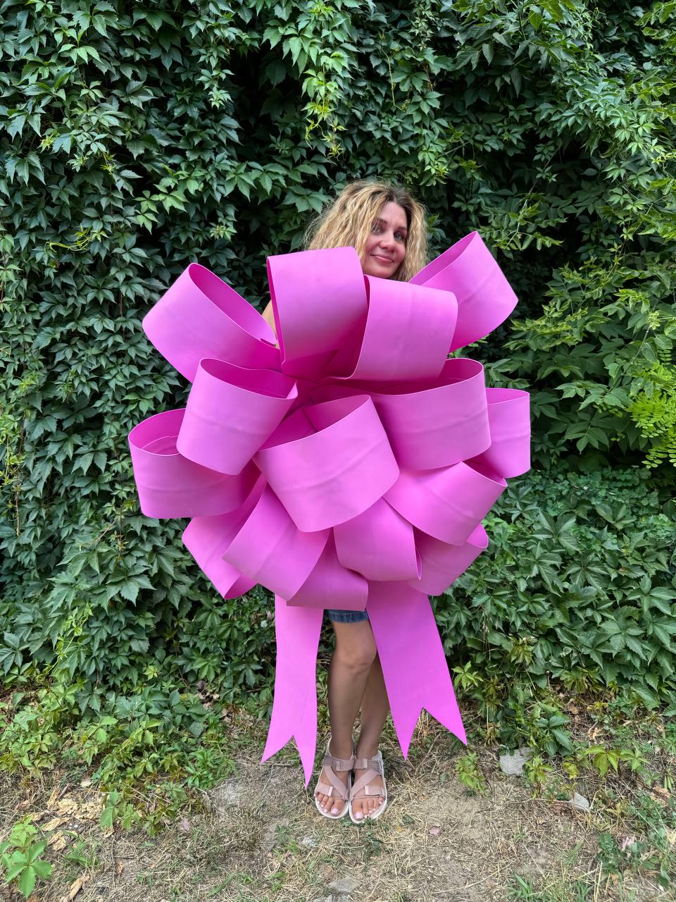 May include: A large pink bow made of multiple layers of ribbon. The bow is large enough to cover a person's torso. The bow is tied with a long ribbon that trails behind the person.