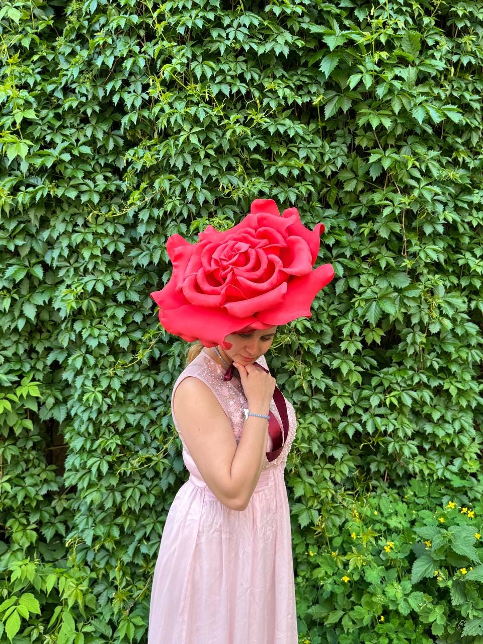 May include: A woman wearing a large, red rose-shaped hat and a light pink dress. The hat is the focal point, with detailed petals. The background is a wall of green ivy. The dress has a V-neck and a dark red ribbon.