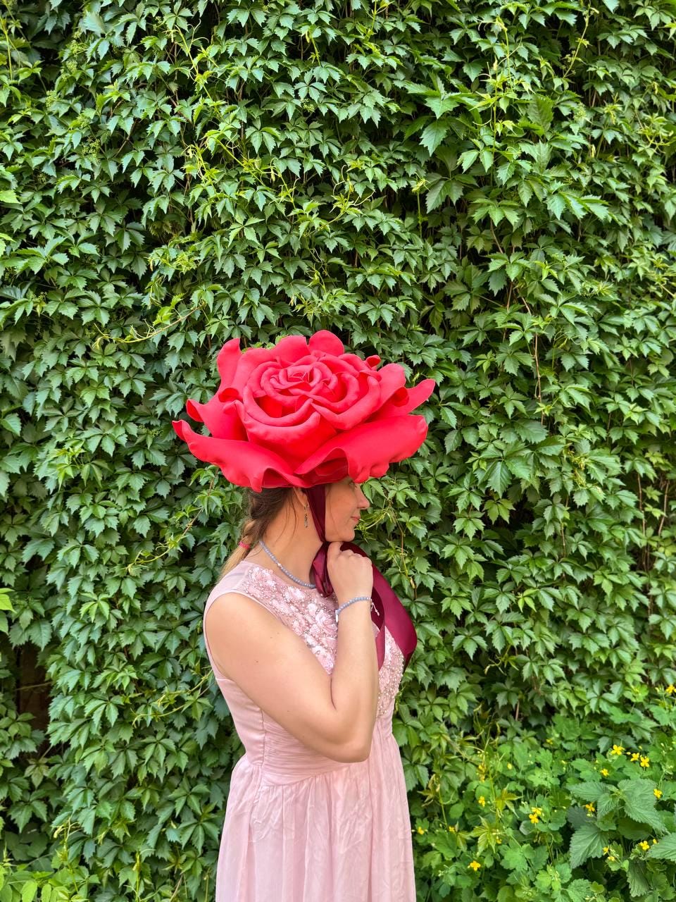 May include: A woman wearing a large, red rose-shaped hat. The hat is the focal point, with its detailed petals and vibrant color. She is wearing a pink dress and standing in front of a wall of green foliage. The hat is a unique accessory.