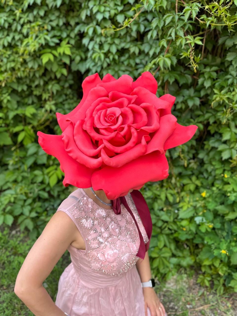 May include: A large, vibrant red rose headpiece. The oversized floral design covers the head, with detailed petals. The person is wearing a pink dress with floral embellishments. The background is a lush green wall of foliage.