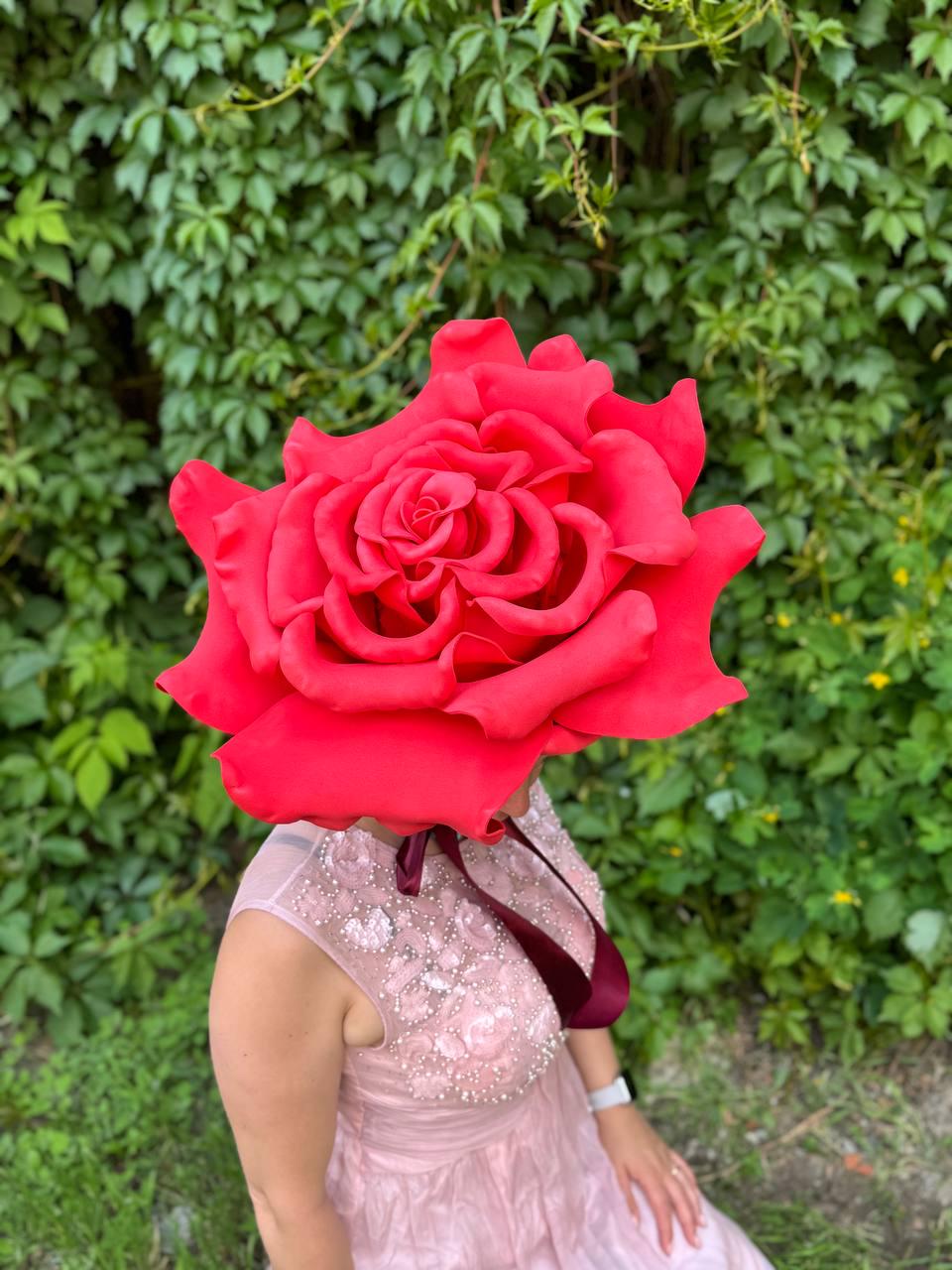 May include: A large, vibrant red rose headpiece. The oversized floral design covers the head, with a burgundy ribbon detail. The person is wearing a light pink dress with beaded embellishments.