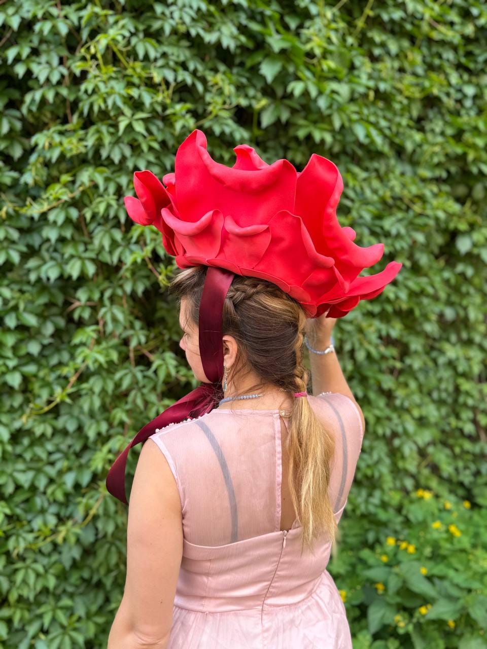 May include: A vibrant red headpiece resembling a large flower, worn by a person with blonde hair. The headpiece is the focal point, with a burgundy ribbon accent. The background is a lush green wall of foliage.