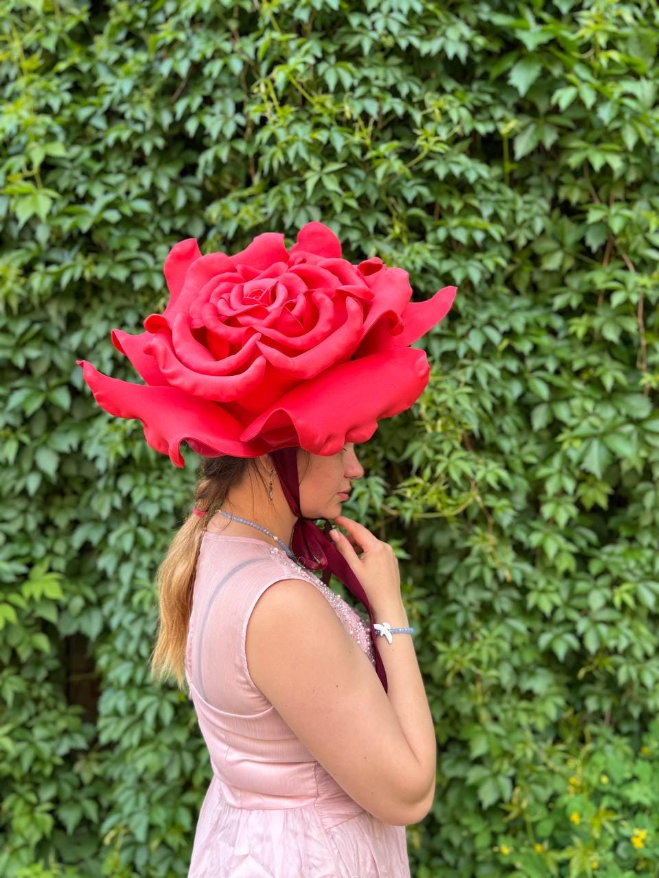 May include: A large, vibrant red rose-shaped hat. The hat is worn by a person in a pink dress, with a green leafy background. The hat's petals are detailed, and the overall effect is striking and whimsical.