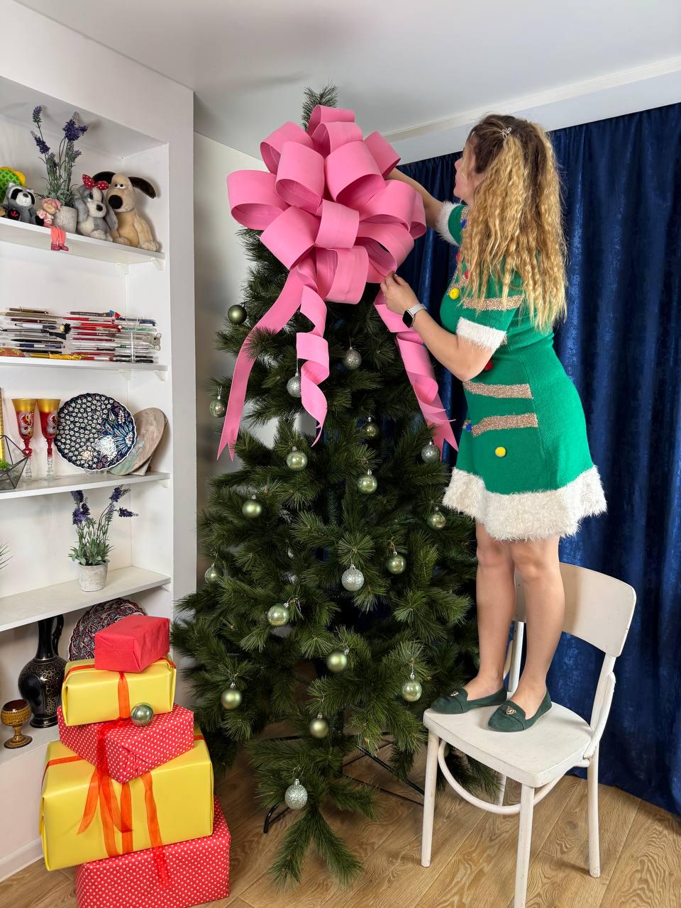 May include: A decorated Christmas tree with a large pink bow on top. The tree is adorned with silver ornaments and surrounded by wrapped gifts in red and yellow. A person is standing on a chair, adjusting the bow.