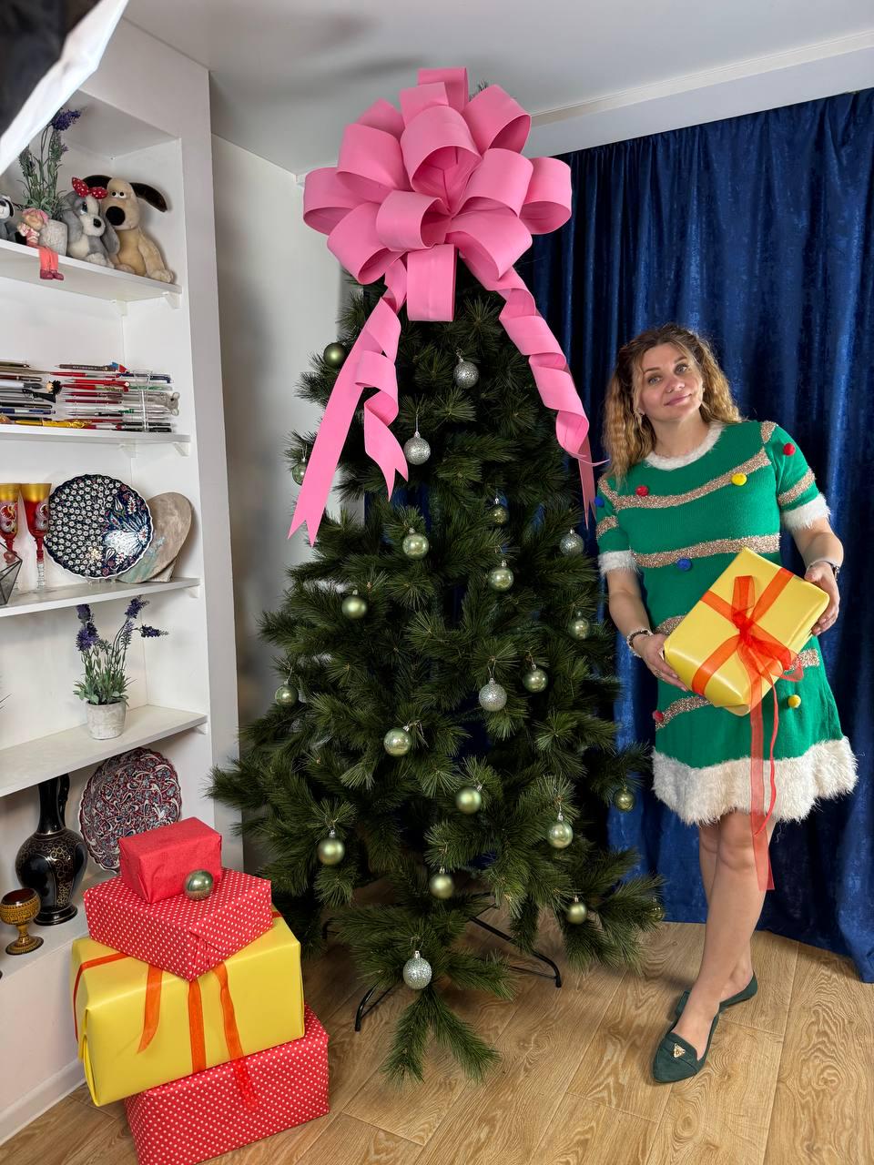 May include: A decorated Christmas tree with a large pink bow on top, silver and green ornaments, and a woman wearing a green dress holding a yellow gift box with a red bow. Several wrapped gifts are stacked on the floor.
